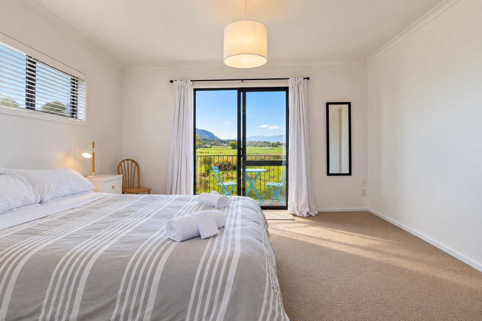 A bedroom with a bed, a chair, a nightstand, a lamp, a window with curtains, and a view of a green landscape.