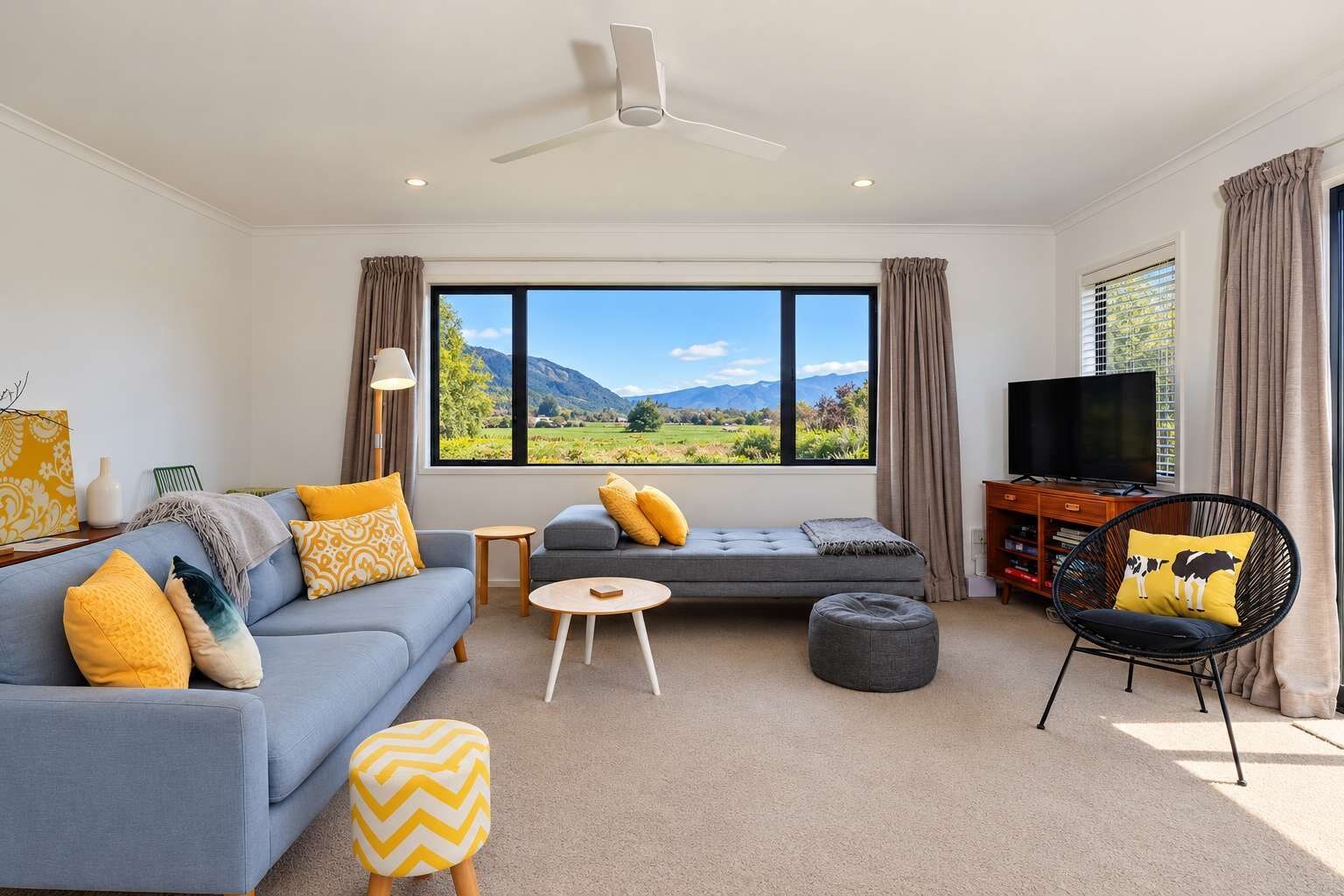 A modern living room with a sofa, ottoman, and a chair, a television, and a large window with a mountain view.