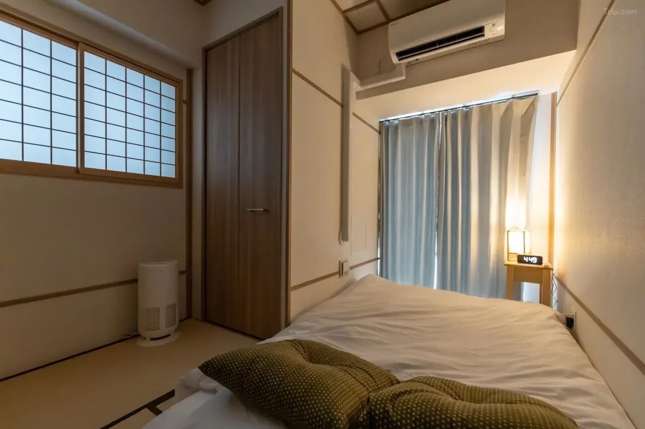 A hotel room with a bed, a window with a grid pattern, an air conditioning unit, a bedside table with a lamp, and a door.