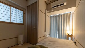 Ein Hotelzimmer mit Bett, Fenster mit Gittermuster, Klimaanlage, Nachttisch mit Lampe und Tür.