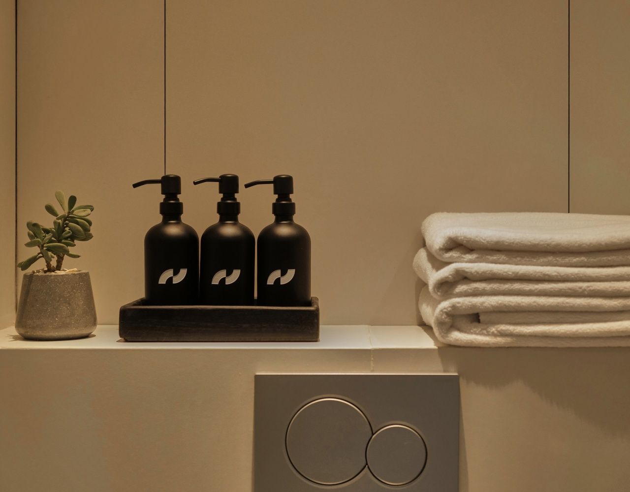 A bathroom with a sink, three black soap dispensers, a potted plant, and a stack of white towels.