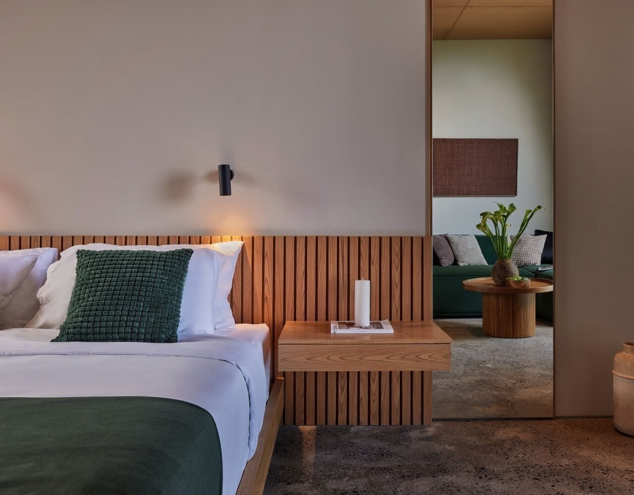 A hotel room with a bed, a nightstand, a mirror, and a view of a green sofa and plant in the adjacent room.