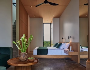 A modern hotel room with a bed, a round wooden table with a vase of flowers, and a view of greenery outside.