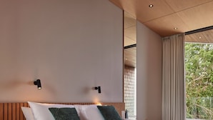 Une chambre d’hôtel moderne avec une tête de lit en bois, un grand lit avec une literie blanche et verte, deux lampes fixées au mur et une vue sur la verdure par la fenêtre.