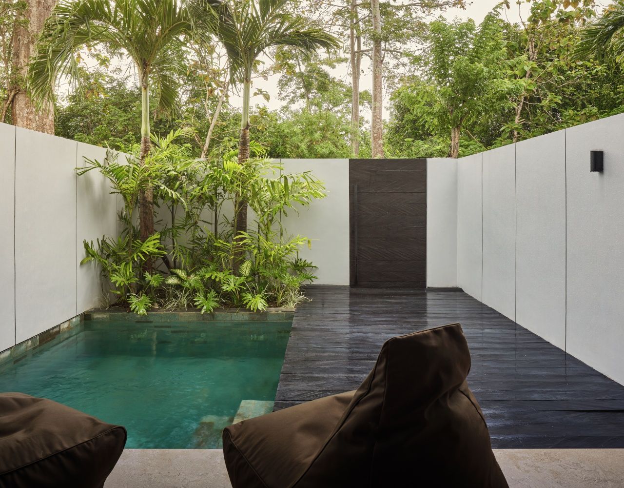 A modern outdoor pool area with a dark floor, surrounded by lush greenery and a white wall.