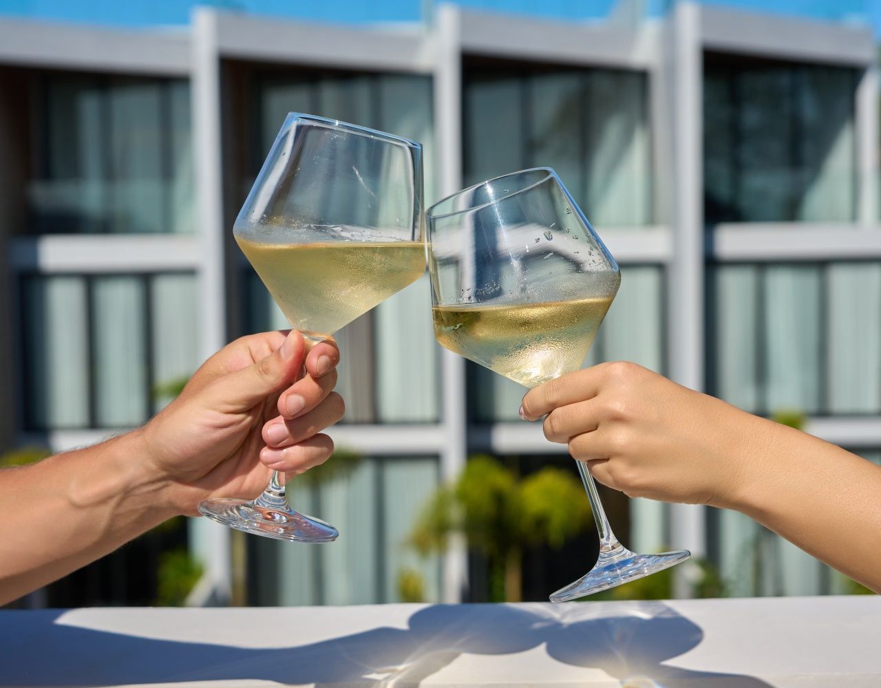Two hands holding wine glasses with a building in the background.