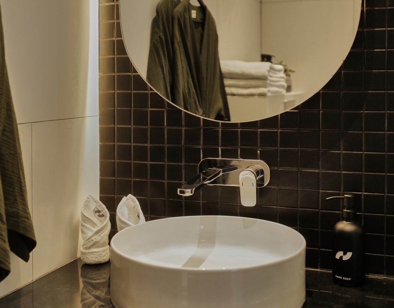 A bathroom with a round white sink, a mirror, and a black tiled wall.