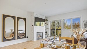Un coin repas avec une table en bois dressée pour quatre personnes, une cheminée et une grande fenêtre donnant sur les arbres.