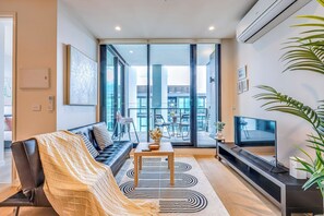 A modern living room with a sofa, a coffee table, a TV, and a large window offering a view of the city. - StayAU Convenient City Apt in West Melb (West Melbourne)