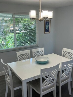 Een eetgedeelte met een witte tafel en stoelen, een kom op de tafel en een raam met jaloezieën.