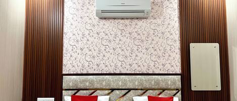 A hotel room with a bed, a wall-mounted air conditioner, a power outlet, and a decorative panel on the wall.