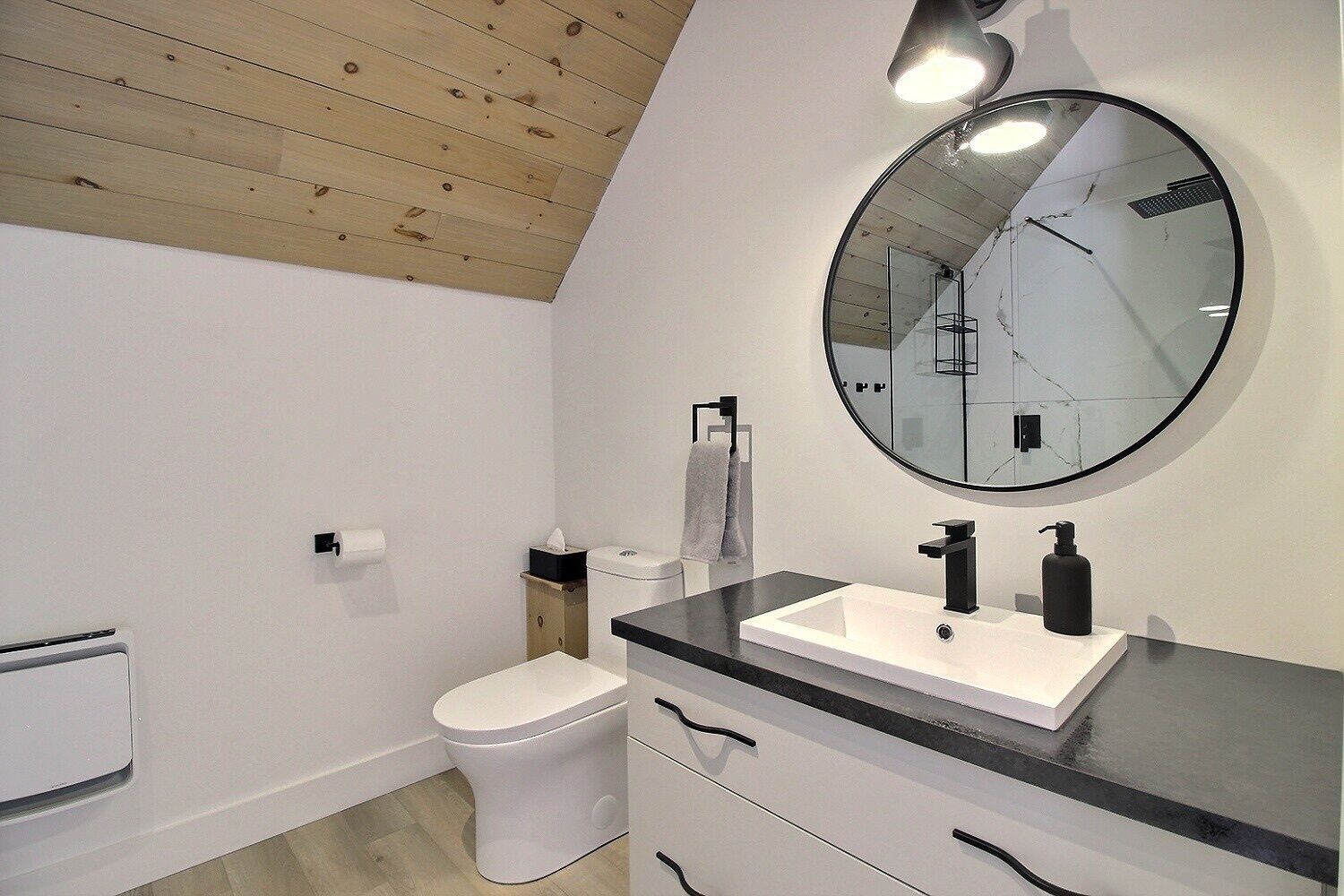 A modern bathroom with a wooden ceiling, a white toilet, a black sink with a large mirror, and a towel rack.
