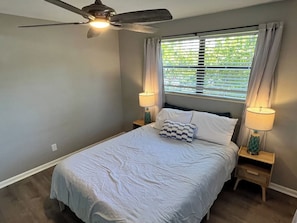A bedroom with a bed, two bedside lamps, a ceiling fan, and a window with curtains. - Ashota - The Driftwood (Sarasota)