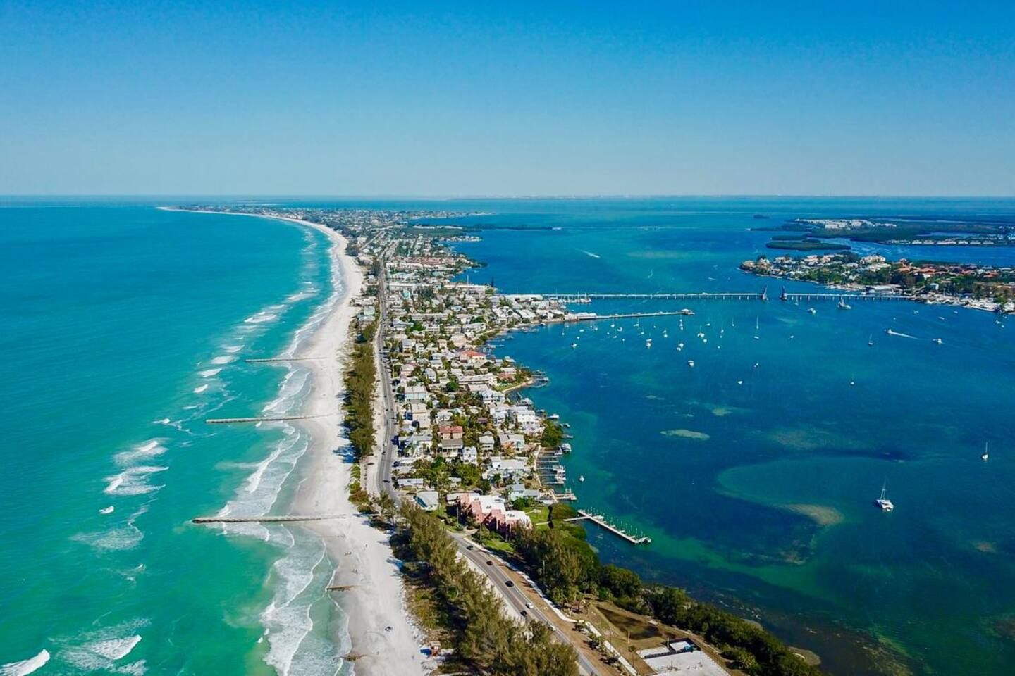 An aerial view of a coastal town with a long beachfront, clear blue waters, and a bridge connecting to an island.