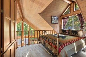 A bedroom with a wooden ceiling, a bed with a patterned bedspread, a window with curtains, and a view of greenery outside.
