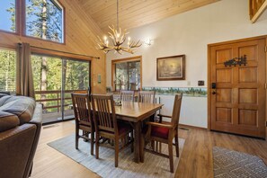 A dining area with a wooden table and chairs, a chandelier, and a view of the outdoors through large windows.