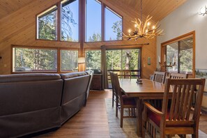 A wooden interior with a dining table, chairs, a sofa, and large windows.