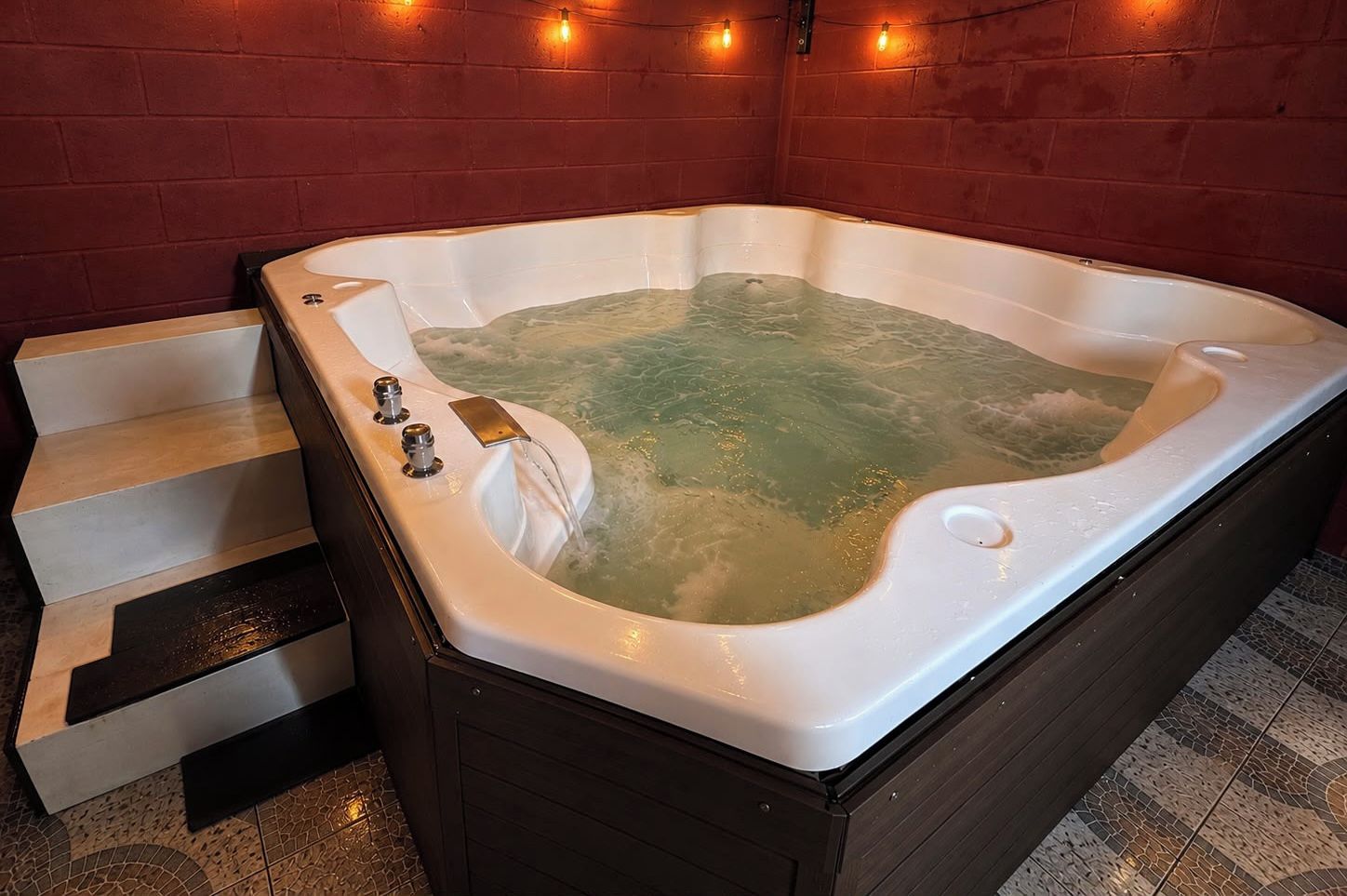 A hot tub with a built-in bench and steps leading into it, set against a brick wall with string lights.