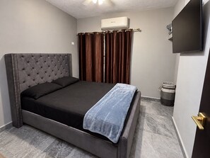 A bedroom with a tufted headboard bed, a flat-screen TV mounted on the wall, a window with brown curtains, and a small trash bin. - Casa Pacifico Azul (Puerto Vallarta) (Ixtapa)