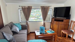 Un salón con sofá, sillón, mesa de centro, televisor y vista panorámica de la ciudad a través de una ventana.