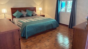 Un dormitorio con piso de madera, una cama con colcha azul, dos lámparas de noche y un ventanal con cortinas.