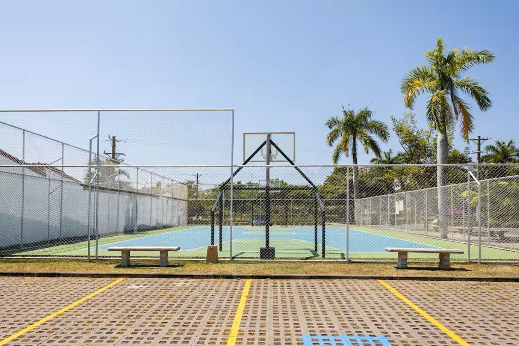An outdoor basketball court with a fence, benches, and palm trees in the background.