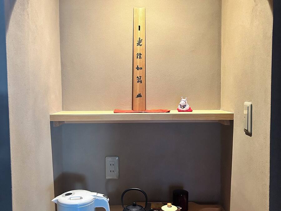 A small wooden shelf with a calligraphy piece, a white teapot, a black teapot, and a red mat.