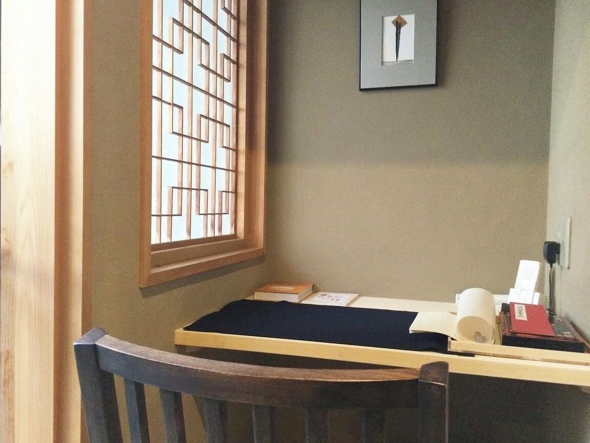 Een werkhoek met een bureau, stoel, raam met jaloezieën en een ingelijst beeld aan de muur.