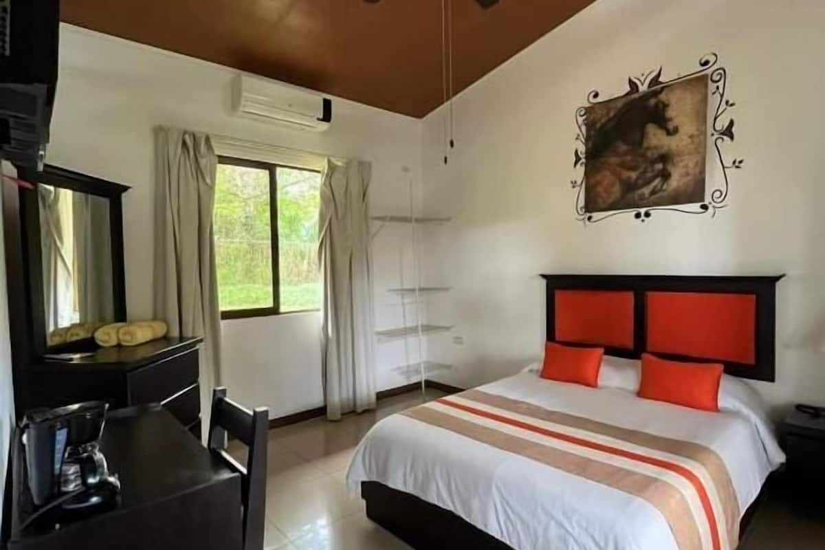 A hotel room with a bed, a desk, a chair, a ceiling fan, a window with curtains, and a painting on the wall.