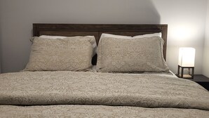 Un llit ben fet amb un llençol i coixins estampats amb motius florals, un panell de fusta i una tauleta de nit amb una làmpada.