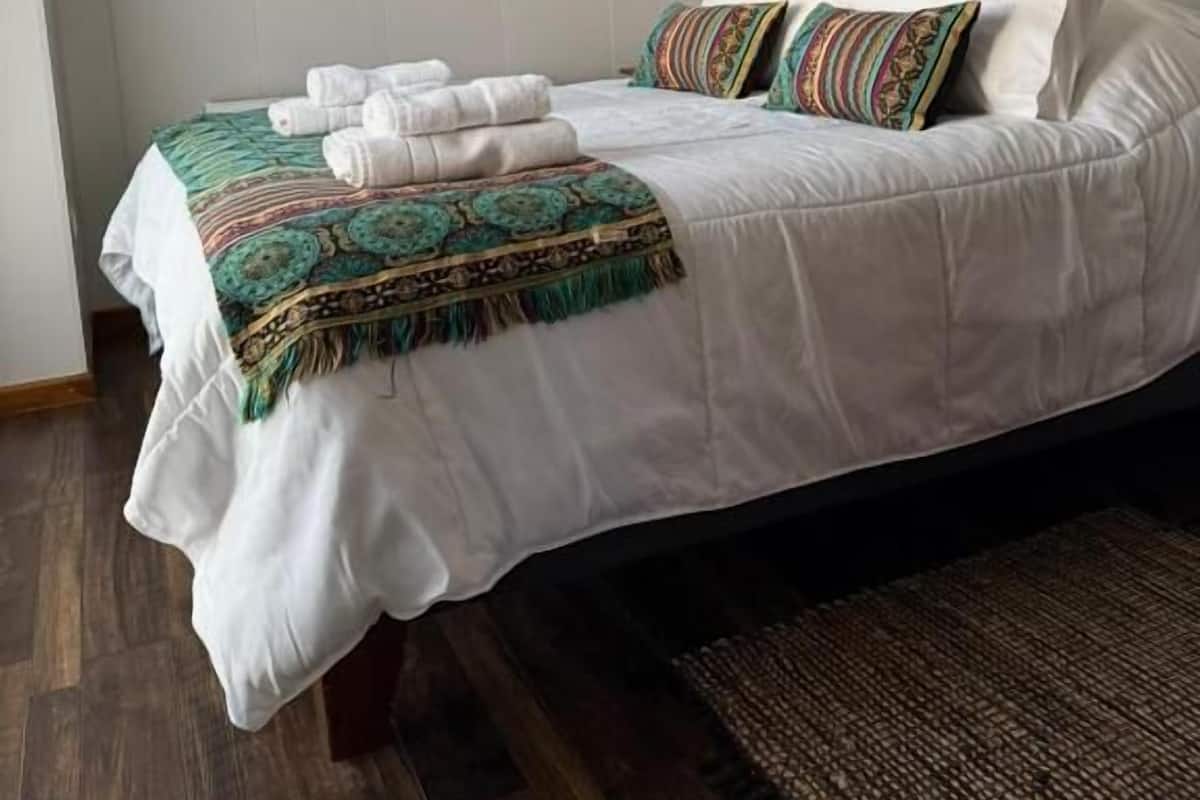 A neatly made bed with white linens and decorative pillows, a window with blinds, and a wooden floor.