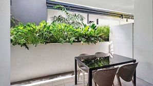 Un área de comedor al aire libre moderna, con mesa y sillas de vidrio, rodeada de plantas verdes exuberantes en una maceta blanca.