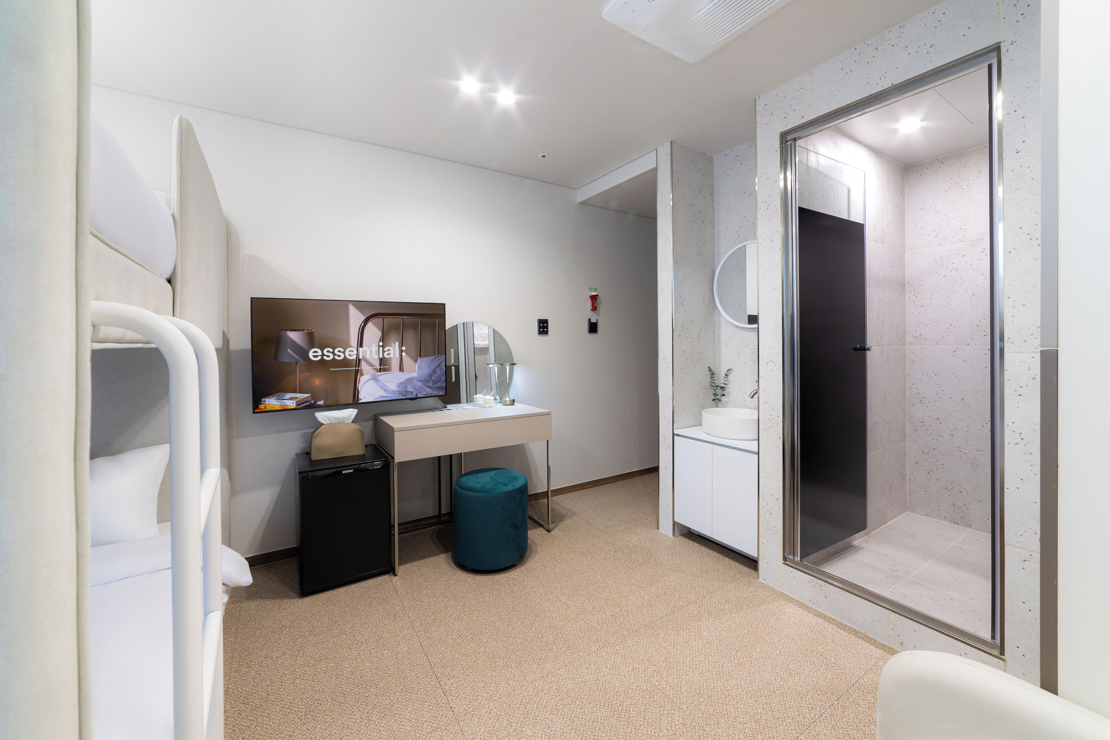 Una habitación de hotel moderna con una cama, un escritorio con computadora, televisor, un cesto de basura pequeño y zona de ducha.