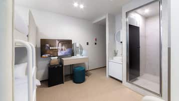 Una habitación de hotel moderna con una cama, un escritorio con computadora, televisor, un cesto de basura pequeño y zona de ducha.