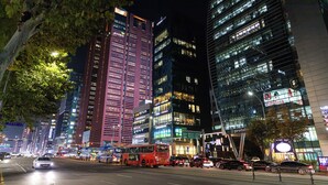 A nighttime cityscape with tall buildings, a red bus, and various vehicles on the street. - Lazy Tiger Hotel (Seoul)