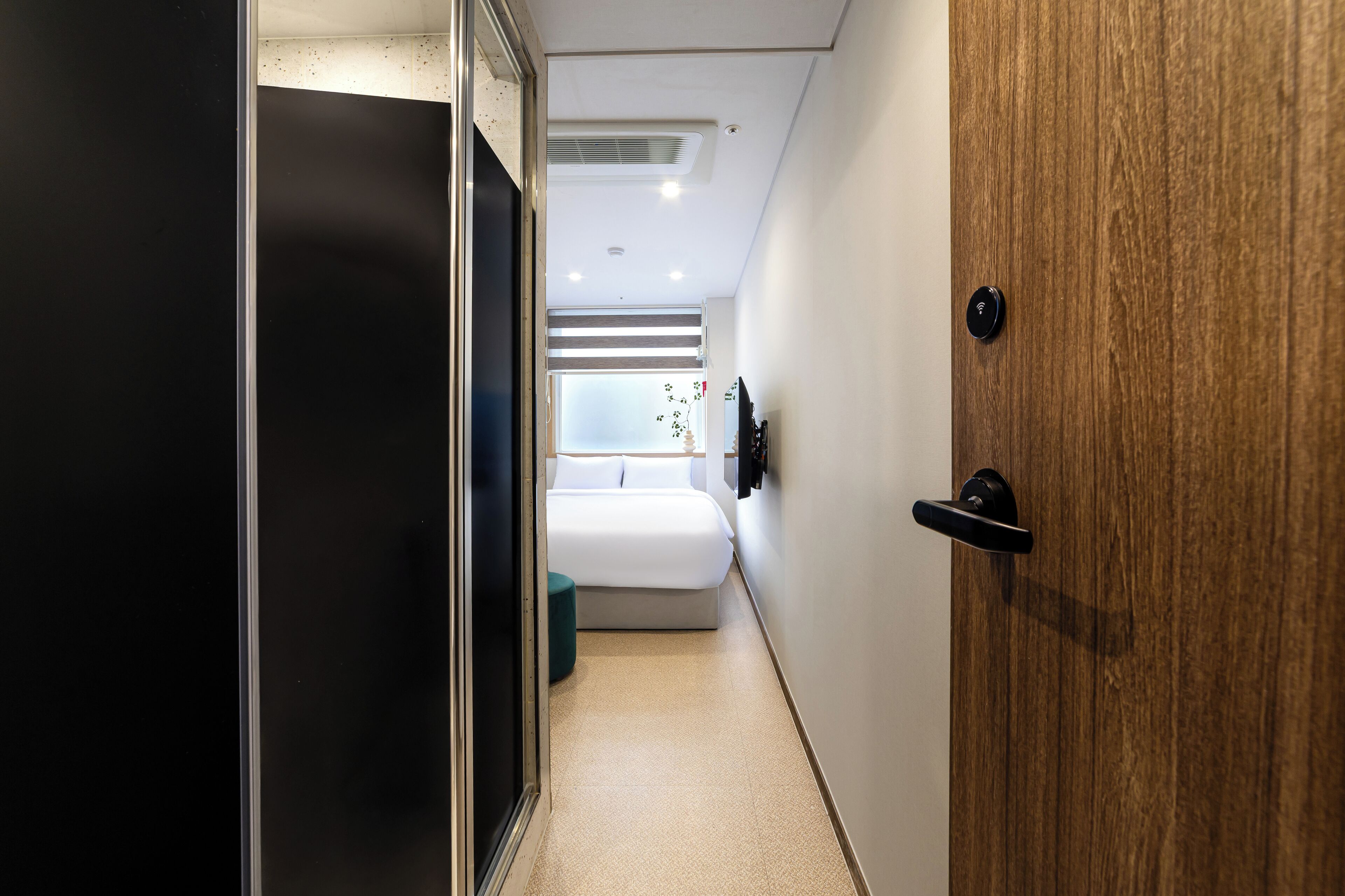Un estrecho pasillo que conduce a un dormitorio con una cama, una puerta de madera y un pequeño ventanal.
