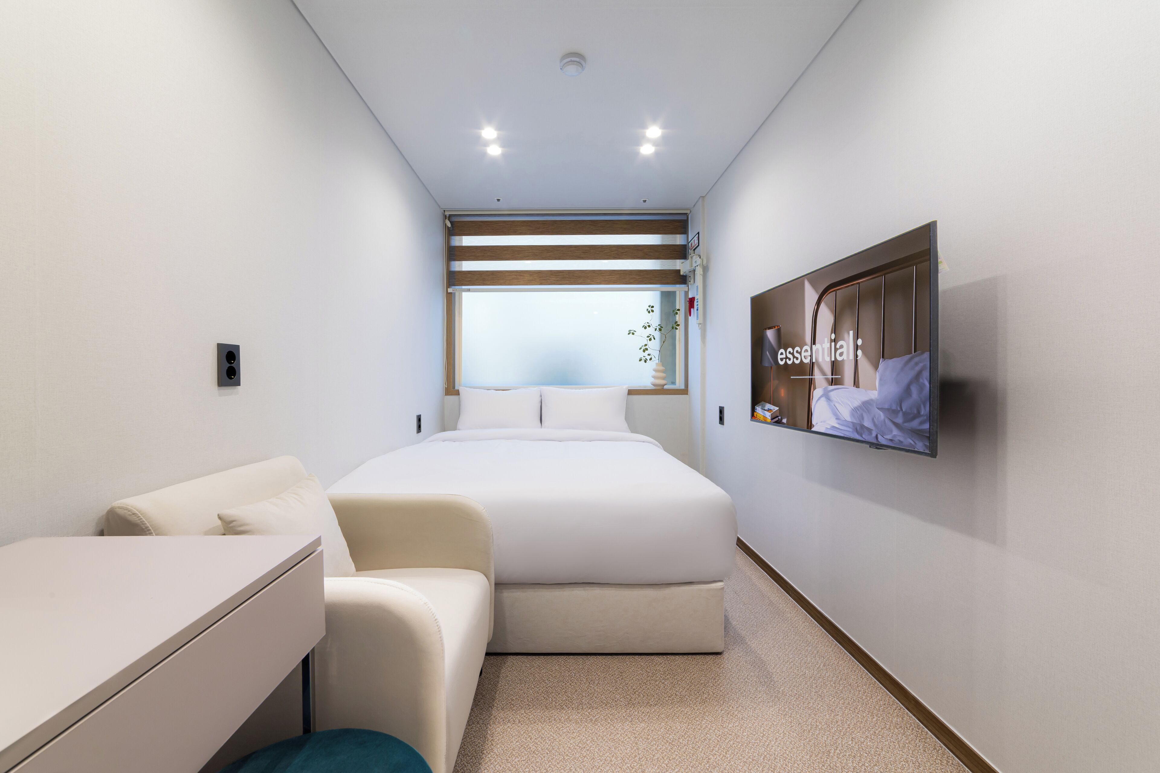 Ein modernes Hotelzimmer mit einem Bett, einem Schreibtisch, einem Stuhl, einem an der Wand montierten Fernseher und einem Fenster mit Jalousien.