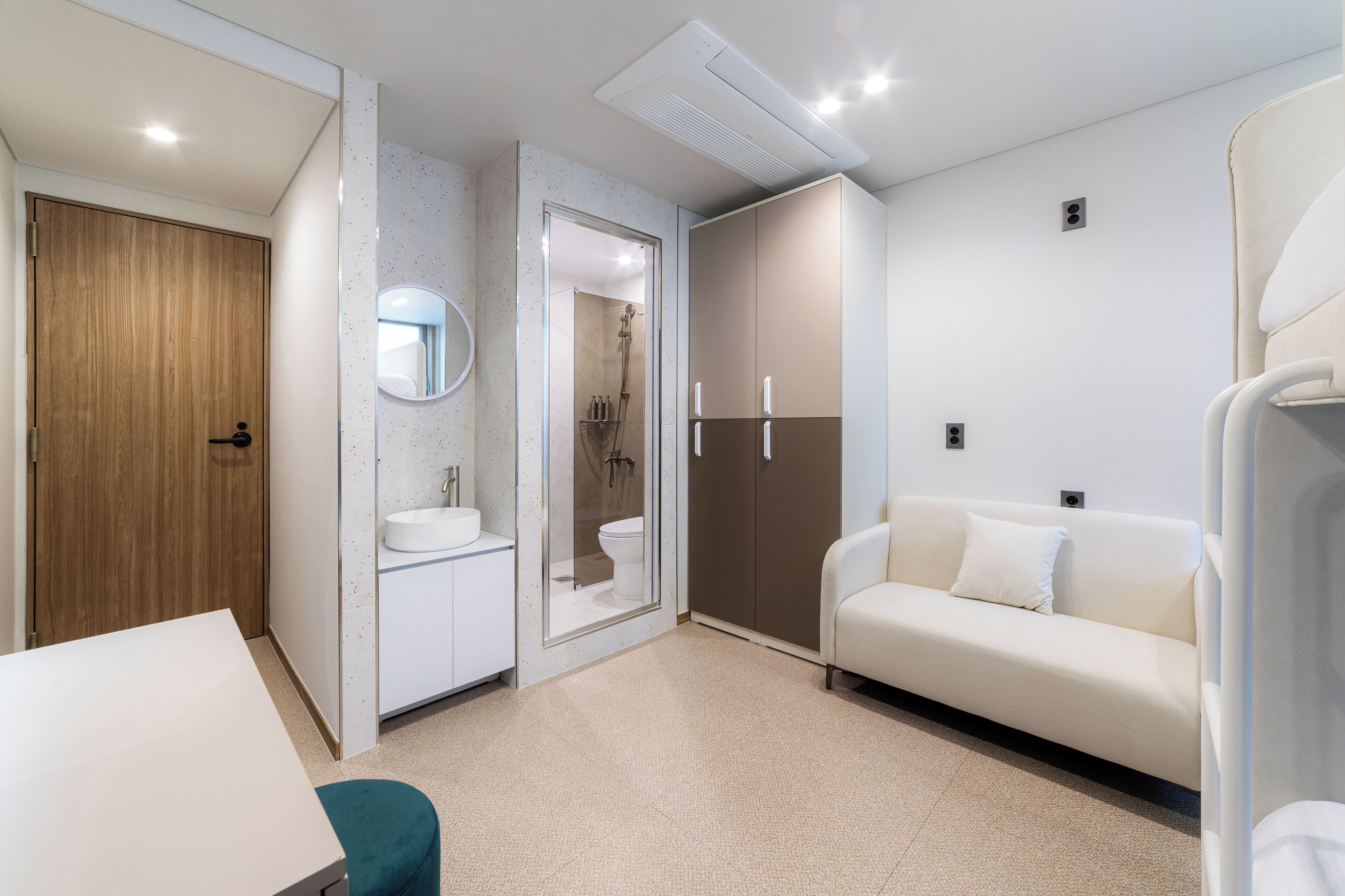 Un baño moderno con puerta de madera, un lavamanos blanco, una ducha con mampara de vidrio y un sofá color beige.