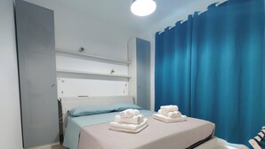 Ett hotellrum med en säng, ett nattduksbord, en hylla med en liten växt och ett fönster med blå gardiner.