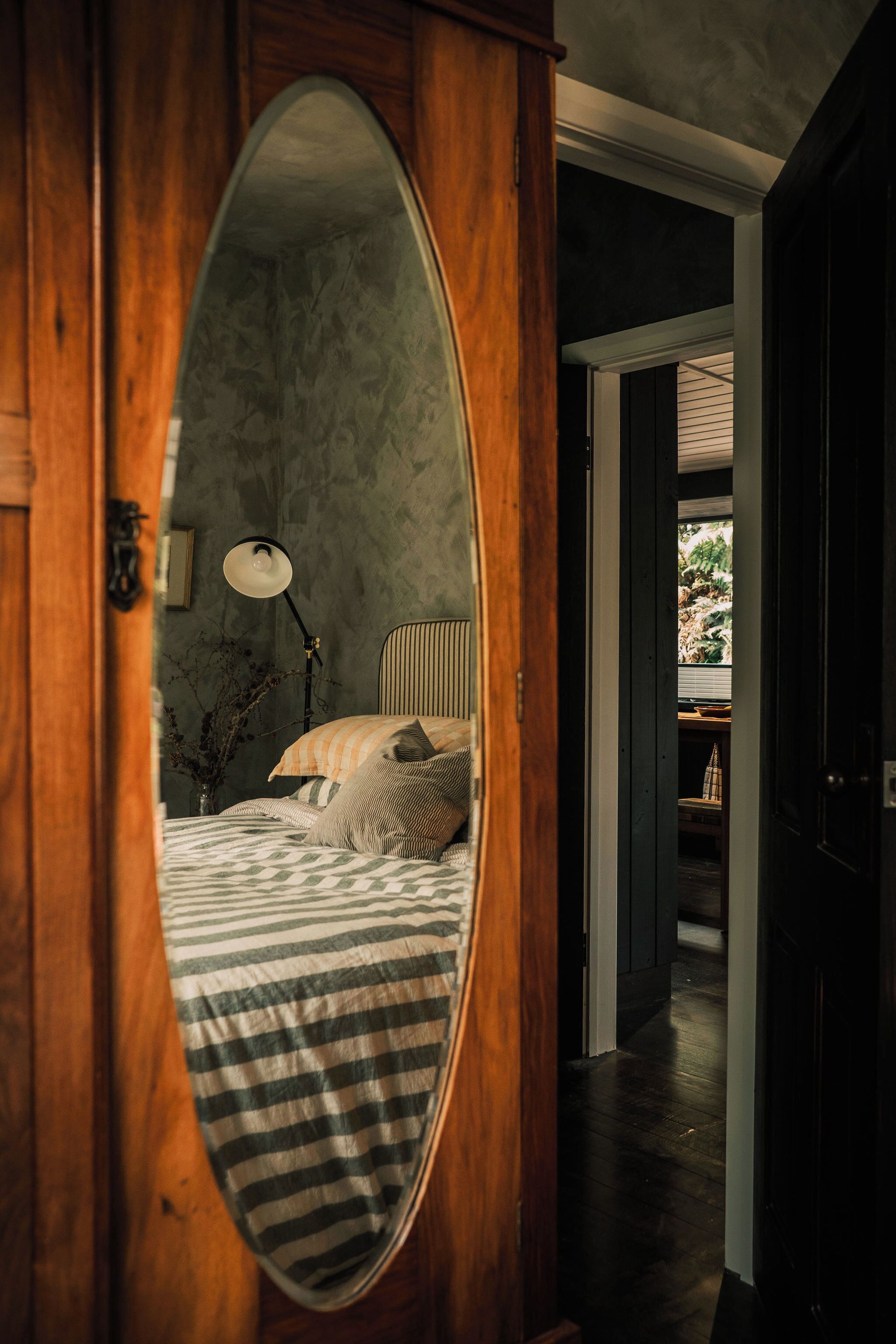 A bedroom with a large mirror, a bed with striped bedding, a bedside lamp, and a view of a garden through a window.