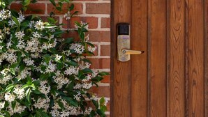 A wooden door with a lock, a brick wall with a number plaque, and a plant with white flowers. - Relax, Explore, Unwind — Coastal Comfort at The Little Monaco (Nelson)