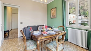A dining area with a wooden table set for four, a sofa, and a view of a mountain through a window. - La Caravella: Stresa Lake & Islands View Balcony (Stresa)