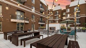 Un patio al aire libre con bancos y mesas de madera, luces colgantes y plantas en macetas.