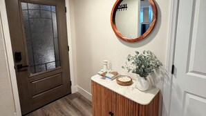 Un pasillo con puerta de madera, un espejo, un mueble pequeño con adornos y una planta en maceta.