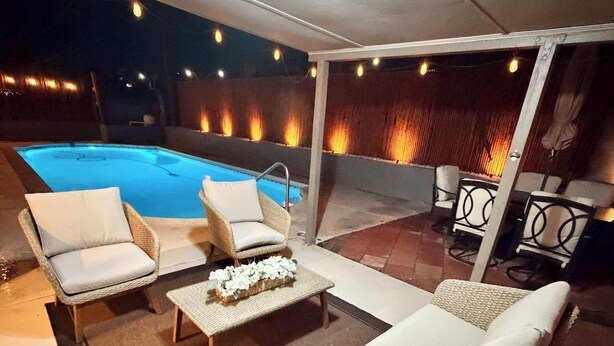 Un patio al aire libre con mobiliario de mimbre, una piscina y luces colgantes.