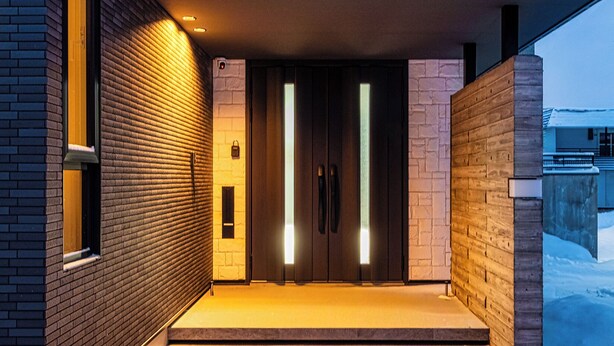 A modern building entrance with a brick facade, a set of steps leading to a dark door, and a snow-covered ground. - THE VELLMORE (Asahikawa)