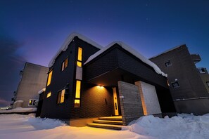 A modern two-story building with a snow-covered roof and a lit entrance. - THE VELLMORE (Asahikawa)