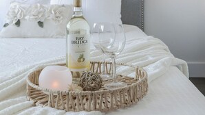 A neatly made bed with a bottle of Bay Bridge wine, a glass, and a decorative basket. - Lux New House by UA/Train/Rogers Place/Hospital/Ravine/AC/D-garage/sleep 12 (Edmonton)