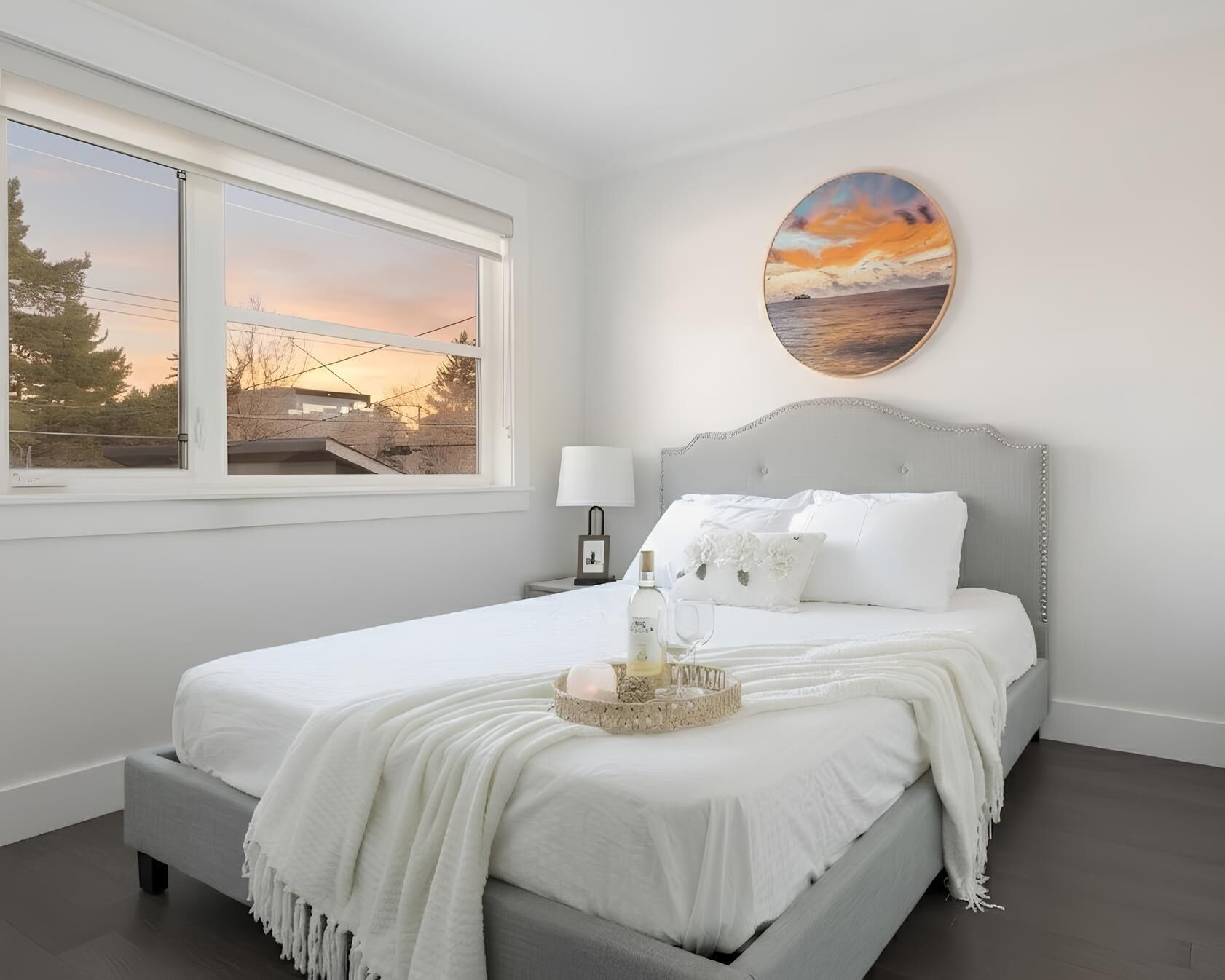 A bedroom with a bed, a nightstand, a lamp, a painting, and a view of trees and a sunset through a window.