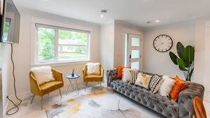 A modern living room with a grey sofa, two yellow armchairs, and a large window offering a view of greenery. - Lux New House by UA/Train/Rogers Place/Hospital/Ravine/AC/D-garage/sleep 12 (Edmonton)